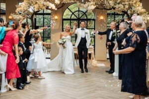 A couple in wedding attire walks hand in hand down a wooden aisle as guests throw white petals in the air, celebrating their confetti exit at Woodlands Hotel, Leeds