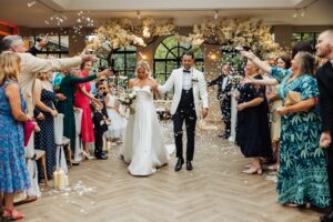 A couple in wedding attire walks hand in hand through a room filled with guests throwing confetti