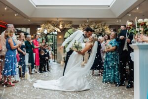 A couple in wedding attire share a kiss as confetti falls around them, with guests clapping and watching in the background at Woodlands Hotel, Leeds