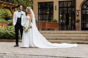 A couple in wedding attire stands close together, smiling at each other, on a paved area in front of a building with large windows