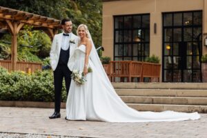 A couple in wedding attire stands together on a paved area in front of a building with large windows at Woodlands Hotel, Leeds