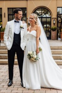 A couple in wedding attire stands together on a paved area in front of a building with large windows and a door at Woodlands Hotel, Leeds