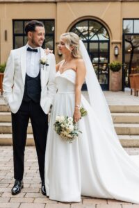 A couple in wedding attire stands together on a paved area in front of a building with arched windows at Woodlands Hotel, Leeds