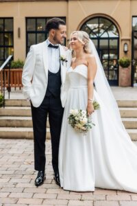 A couple in wedding attire stands close together on a paved area in front of a building with large windows at Woodlands Hotel, Leeds