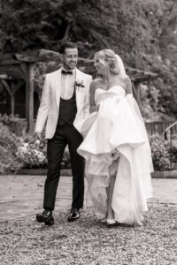 A couple in wedding attire walks together on a gravel path, smiling and holding a bouquet at Woodlands Hotel, Leeds