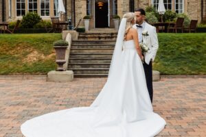 A couple in wedding attire stands close together on a paved area in front of a stone staircase leading to a building with large windows at Woodlands Hotel, Leeds