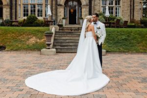 A couple in wedding attire stands on a brick-paved area in front of a stone staircase leading to a building with large windows at Woodlands Hotel, Leeds