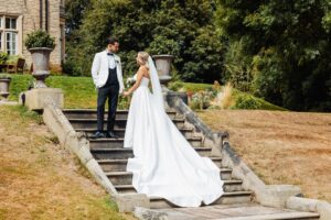 A couple in wedding attire stands on a stone staircase, holding hands and looking at each other at Woodlands Hotel, Leeds