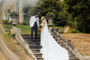 A couple in wedding attire stands on a stone staircase, holding hands and smiling at each other at Woodlands Hotel, Leeds