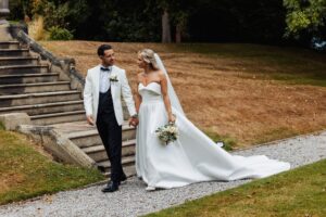 A couple in wedding attire walks down a stone staircase, the bride holding a bouquet and the groom in a white tuxedo