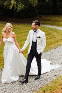 A couple in wedding attire holds hands and smiles at each other on a gravel path surrounded by grass and trees at Woodlands Hotel, Leeds