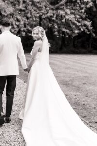 A bride in a white wedding dress and a man in a dark suit walk hand-in-hand in an outdoor setting with trees in the background