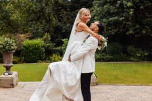 A couple in wedding attire embrace on a paved area with greenery and a stone planter in the background at Woodlands Hotel, Leeds
