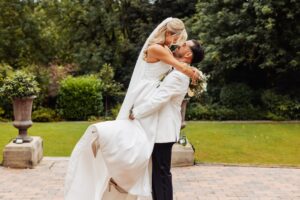 A couple in wedding attire embraces outdoors in a garden setting with trees in the background at Woodlands Hotel, Leeds