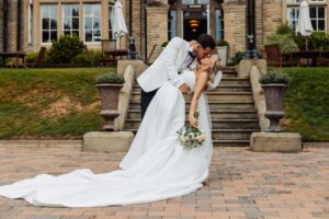 A couple in wedding attire share a kiss on a brick-paved area in front of a grand building with stone steps and large windows