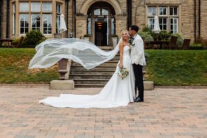 A couple in wedding attire stands in front of a stone building with a flowing veil, on a paved area with a stone staircase and outdoor seating