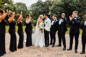 A bride in a white gown and a groom in a white tuxedo kiss, surrounded by bridesmaids in black dresses and groomsmen in black suits at Woodlands Hotel, Leeds