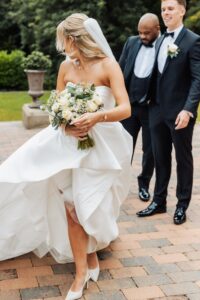 A bride in a strapless white wedding dress holds a bouquet of white roses and greenery while walking on a paved path with a man at Woodlands Hotel, Leeds