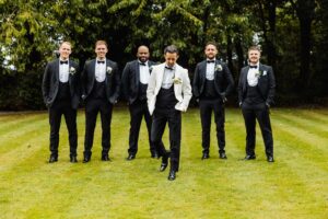 A group of men in formal attire, including tuxedos and a white suit, stand on a grassy lawn with trees in the background at Woodlands Hotel, Leeds