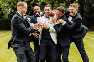 A group of men in formal attire, including tuxedos and white shirts, are gathered outdoors on a grassy area, laughing and celebrating at Woodlands Hotel, Leeds