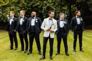 A group of men in formal attire, including a white tuxedo and black tuxedos, stand on a grassy area at Woodlands Hotel, Leeds