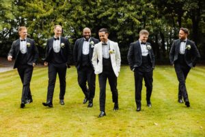 A group of men in formal attire, including tuxedos and a white blazer, walk on a grassy area, smiling and wearing boutonnieres