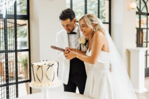 A couple in wedding attire cuts a white cake with black ribbon decorations, standing indoors with large windows and modern interior design at Woodlands Hotel, Leeds