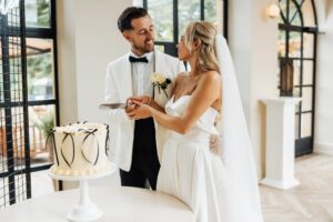 A couple in wedding attire stands together, holding a knife to cut a cake decorated with white frosting and black ribbons at Woodlands Hotel, Leeds