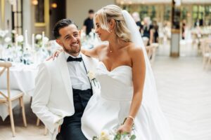 A couple in wedding attire stands close together in a warmly lit room with tables and candles in the background