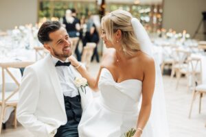 A couple in wedding attire is seated together in a warmly lit room with wooden flooring and tables set for a meal at Woodlands Hotel, Leeds
