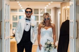 A couple in wedding attire stands in a doorway, the man in a white tuxedo and the woman in a strapless white dress at Woodlands Hotel, Leeds
