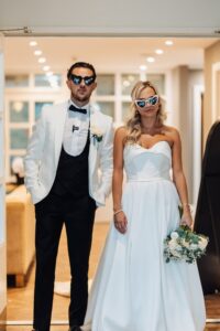 A couple in wedding attire, the man in a white tuxedo and the woman in a strapless white gown, both wearing sunglasses with text on them
