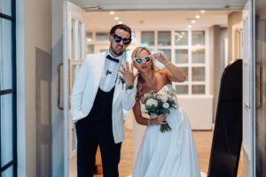 A couple in wedding attire stands in a doorway, the man in a white tuxedo and the woman in a white wedding dress holding a bouquet at Woodlands Hotel, Leeds