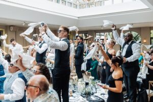 A group of people raise white napkins in the air at a wedding reception, dressed in formal attire, with a festive and celebratory atmosphere