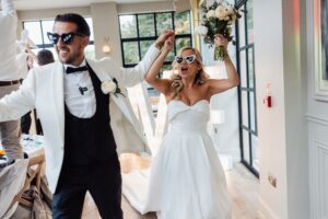 A couple in wedding attire dances joyfully in a well-lit room with large windows and wooden flooring