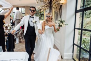 A couple in wedding attire walks hand in hand through a warmly lit room with large windows and greenery outside at Woodlands Hotel, Leeds