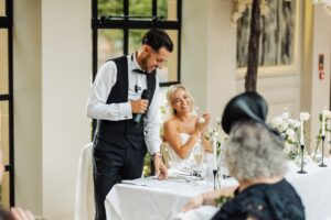 A man in a dark suit and bow tie delivers a speech at a wedding reception, with a woman in a white dress clapping and smiling beside him at Woodlands Hotel, Leeds