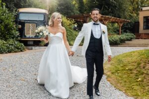 A couple in wedding attire walks hand in hand on a gravel path, with a vintage car and wooden pergola in the background