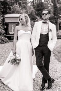A couple in wedding attire walks together on a gravel path, with a vintage car and trees in the background
