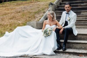 A couple in wedding attire is seated on a stone staircase, with the woman in a strapless white gown and holding a bouquet of white flowers at Woodlands Hotel, Leeds
