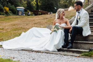 A couple in wedding attire sits on a stone staircase, the woman holding a bouquet of white flowers