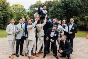 A group of men in formal attire hold another man in the air, celebrating outdoors in a garden setting at Woodlands Hotel, Leeds