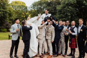 A group of men in formal attire lifts a woman in a white wedding dress, celebrating outdoors on a sunny day at Woodlands Hotel, Leeds