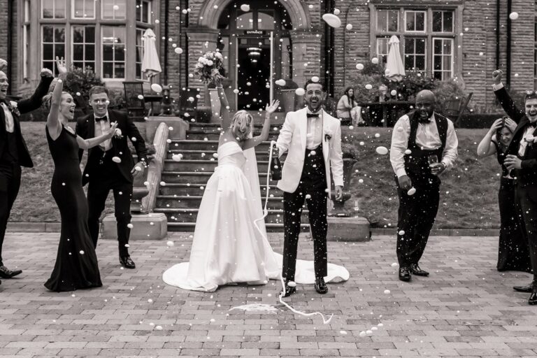 Confetti falls as the bride and groom are surrounded by guests in formal attire, celebrating their wedding exit at Woodlands Hotel, Leeds