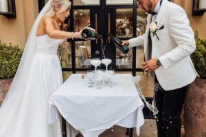 A bride and groom pour champagne into a pyramid of glasses, creating a festive scene outdoors at Woodlands Hotel, Leeds