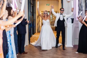 A bride and groom walk hand in hand down a hallway, with guests raising white sparklers on either side, creating a festive atmosphere at Woodlands Hotel, Leeds