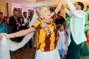 A bride in a white gown and a maroon and yellow striped football shirt dances joyfully with a man in a light blue suit at Woodlands Hotel, Leeds