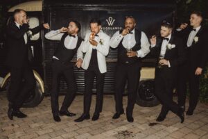 A group of men in formal attire, including tuxedos and suits, are standing in front of a vintage vehicle, smoking and holding drinks at Woodlands Hotel, Leeds