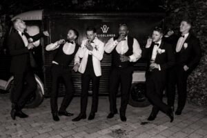 A group of men in formal attire, including tuxedos and white shirts, are standing in front of a vintage car at Woodlands Hotel, Leeds