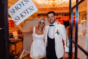 A couple in wedding attire stands close together in a warmly lit room, with one holding a sign that reads 'SHOTS!